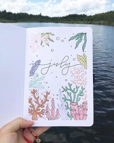 someone holding up an open notebook with the word july written on it in front of a lake