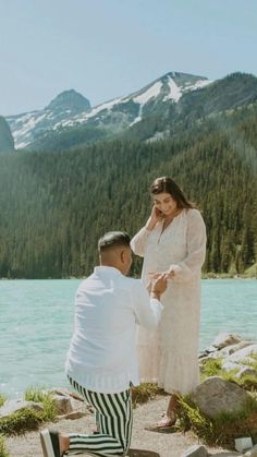 a man kneeling down next to a woman on the shore of a lake with mountains in the background