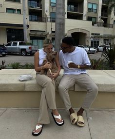 a man and woman sitting on a ledge looking at their cell phones while holding a dog