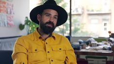 a man with a beard wearing a yellow shirt and black hat sitting in front of a table