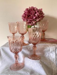 four pink wine glasses sitting on top of a white table cloth next to a vase with purple flowers in it