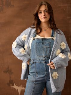 a woman wearing overalls and a sweater with flowers on the sleeves is standing in front of a brown wall