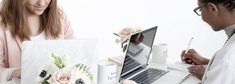 two women sitting at a table working on their laptops and one is holding a pen