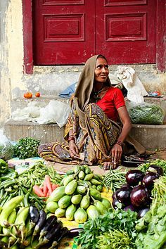 a woman sitting on the ground surrounded by fruits and vegetables in front of a red door