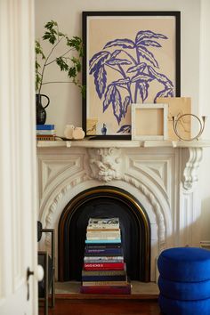 a fireplace with books stacked on top of it in front of a blue ottoman and framed artwork