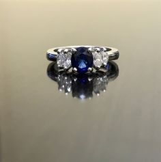 a three stone ring with an oval blue sapphire surrounded by round brilliant cut diamonds on a reflective surface