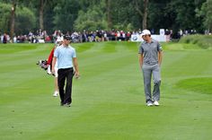 two men walking across a green covered golf course with people watching from the sidelines