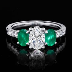 an emerald and diamond three stone ring on a black surface with reflection in the middle