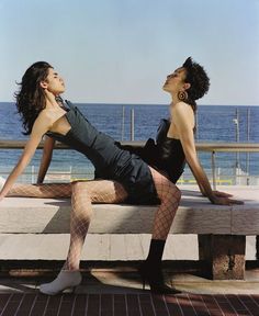 two women are sitting on a bench near the ocean and one is wearing fishnet stockings