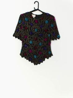 Vintage sequin top in black with floral pattern by Laurence Kazar. This beautiful floral sequin and beaded top features bright blue, pink and purple flowers, short sleeves, shoulder pads, a quarter zip and hook and eye closure on the back. Made from a silk and a polyester lining, this top is the perfect party piece! Our recommended size: Medium Label says: Large Condition: Very good* Material: 100% silk Lining material: 100% polyester  Measurements in inches: Pit to pit: 19 Shoulders: 16 Sleeve: Fitted Sequin T-shirt For Summer, Black Spring Top With Contrast Sequin, Spring Black Tops With Contrast Sequin, Spring Black Top With Contrast Sequin, Black Tops With Contrast Sequin For Spring, Black Sequin Top For Spring, Fitted T-shirt For Party In Spring, Summer Purple Sequined Tops, Elegant Fitted Multicolor Tops