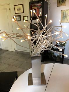 a white table topped with a metal vase filled with branches covered in fairy lights next to a bookshelf