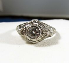 This is a gorgeous platinum antique ring that features a beautiful central diamond. The design of this ring is outstanding. It has very detailed filigree and carving and engraving goes halfway down the band. The diamond is a bezel set brilliant cut diamond and weighs .57 carats. It is a very clean VS-1 and G color. It has a ton of sparkle! There are 2 more diamonds on the shoulders and 2 on the gallery side. The total weight is estimated at .70 carats. All the diamonds are clean and sparkly. It Vintage Silver Filigree Ring With Diamond Accents, Heirloom Silver Engraved Ring With Center Stone, Antique Engraved Diamond Ring With Accents, Antique White Gold Diamond Cut Ring, Vintage Filigree Ring With Diamond Cut, Heirloom Silver Diamond Ring With Intricate Design, Ornate Filigree Diamond Ring With Brilliant Cut, Ornate Round Diamond Ring With Center Stone, Ornate Brilliant Cut Ring For Anniversary