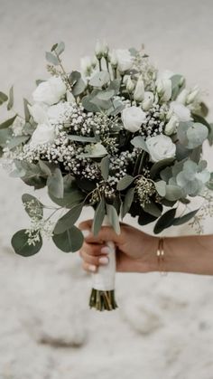 a person holding a bouquet of white flowers and greenery in their hand on the beach