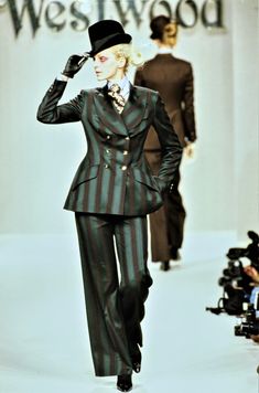 a woman walking down a runway wearing a suit and hat with her hand on her head