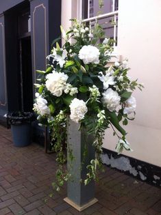 a large vase with white flowers and greenery in front of a building on a brick sidewalk