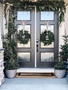 two christmas wreaths on the front door of a house with potted evergreen trees