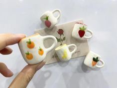 someone is holding some miniature tea cups and spoons on a white surface with fruit painted on them