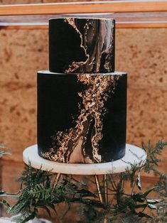 three tiered black and gold marbled cake on a white stand with greenery