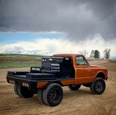 an orange and black truck is parked in the dirt with dark clouds behind it on a cloudy day