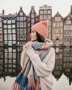 a woman wearing a pink hat and scarf standing in front of water with buildings behind her