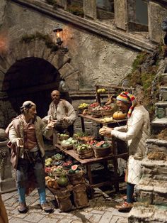 three people standing around a table filled with fruits and veggies in front of an old stone building