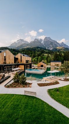 an outdoor swimming pool surrounded by grass and wooden buildings with mountains in the back ground