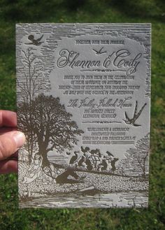 a person holding up a wedding card in front of the camera with trees and people on it