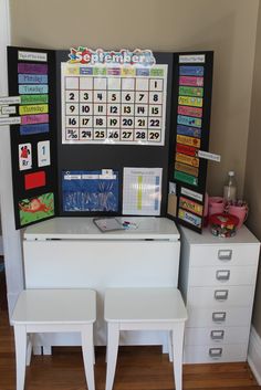 a desk with two white chairs and a calendar on the wall above it, in front of a window