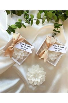 two small white flowers with brown ribbons in boxes on a white satin surface next to greenery