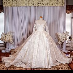 a white wedding dress on display in front of a window with flowers and curtains behind it