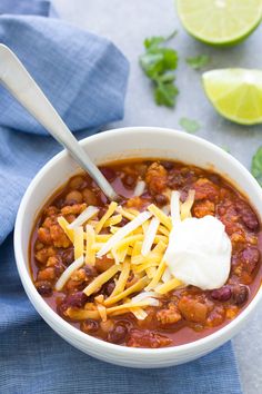 a white bowl filled with chili and cheese next to limes on a blue towel