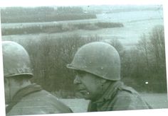 two men wearing helmets are talking to each other in front of a field and trees