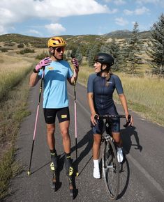 two people riding bikes on the side of a road with skis and poles attached to their backs