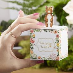 a person holding up a small box with a bear on it's top and flowers around the edges