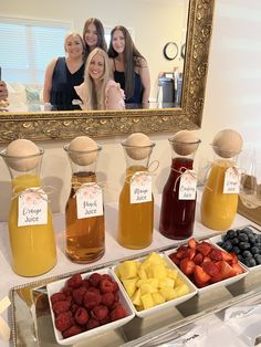 three women standing in front of a table filled with different types of fruit and drinks