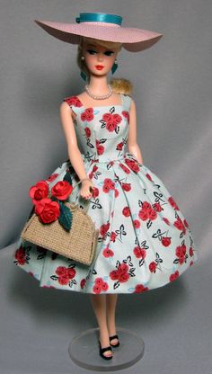 a doll wearing a dress and hat with flowers on it, holding a purse in her hand