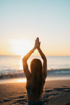 a woman sitting on the beach with her hands up in the air as the sun sets