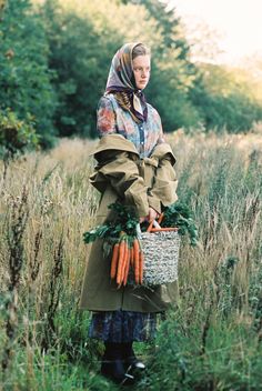 a woman holding a basket full of carrots in a field with tall grass and trees behind her