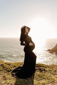 a pregnant woman wearing a black dress and hat standing on the edge of a cliff