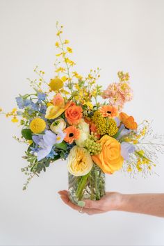 hand holding orange yellow and blue floral arrangement in a glass container in front of a white background. Blue Flower Arrangements, Orange Bouquets, Giving Flowers, Yellow Bouquets, Yellow Wedding Flowers, Best Flowers, Colorful Bouquet, Fresh Flowers Arrangements