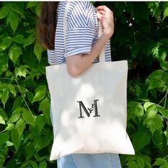 a woman carrying a tote bag with the letter m on it in front of green leaves