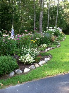 a lush green yard with lots of flowers and rocks on the side of the road