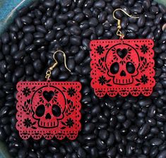 two pairs of red and black sugar skull earrings on top of beans in a bowl