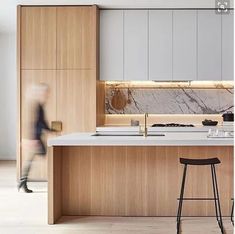 two stools sit in front of an island with marble countertops and cabinets, while a woman walks by