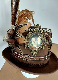 Brown leather steampunk tophat, with peacock feather accents. Perched on the side is a tiny velvet-covered bird, nesting in a flourish of ribbons and feathers. Various bobbles include bullets, charms, beads, and pendants assembled around various ribbons. An antique framed portrait of two children is displayed on the front. All adornments are hand sewn, and the hat is one of a kind. Steampunk Top Hat With High Crown For Themed Events, Steampunk High Crown Hat Band For Festivals, Steampunk High Crown Mini Hat For Themed Events, Vintage High Crown Top Hat For Halloween, Vintage High Crown Top Hat For Costume Party, Vintage Top Hat For Costume Party, Steampunk Mini Hat With High Crown, Vintage High Crown Costume Hats For Themed Events, Vintage Mini Hats With High Crown For Themed Events