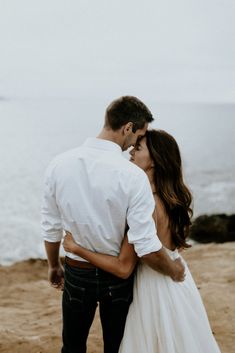 a man and woman standing on top of a sandy beach next to the ocean with their arms around each other