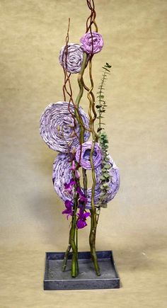 purple flowers are arranged in a vase on a gray base with greenery and branches