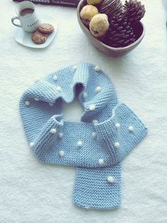 a blue knitted scarf with white pom poms next to a cup of coffee and pine cones