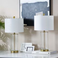 two lamps sitting on top of a white table next to a framed painting and a plant