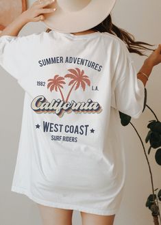 This cool retro California tshirt with palm trees design is the perfect gift idea or souvenir for every surfer in summer. Get yours now! Also available on sweatshirts: https://www.etsy.com/listing/1044743496/california-sweatshirt-los-angeles Browse through my other awesome items here: http://thenims.etsy.com/ UNISEX TEES Gildan 5000™ (CUSTOMER FAVORITE) * 100% Cotton (fiber content may vary for different colors) * Medium fabric (5.3 oz/yd² (180 g/m * Classic fit * Tear away label * Runs true to Retro Text Print T-shirt For Beach, Printed White Tops For Surfing, Retro Surfing T-shirt For Beach Season, White Surfing Shirt With Graphic Print, White Graphic Print Surfing Shirt, Casual Surfing Camp Shirt With Graphic Print, Summer Camp Shirt With Graphic Print For Vacation, Trendy Camp Shirt With Graphic Print For Beach, Summer Graphic Print Camp Shirt For Vacation